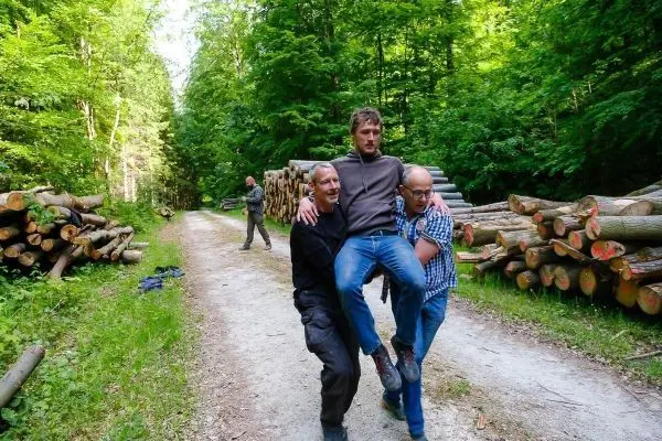 Zwei Personen tragen einen verletzten Mann auf einem Waldweg zwischen Holzstapeln